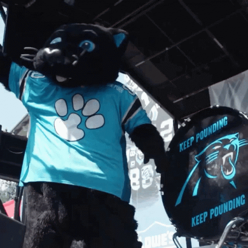 A Black Panther Mascot Wearing A Blue Shirt Stands Next To A Drum That Says Keep Pounding GIF
