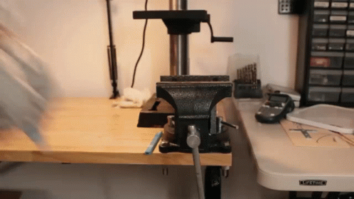 a black vise sits on a wooden workbench next to a drill press