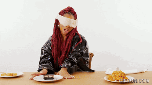 A Blindfolded Woman Sits At A Table With Plates Of Food And A Gierun.com Logo In The Corner GIF