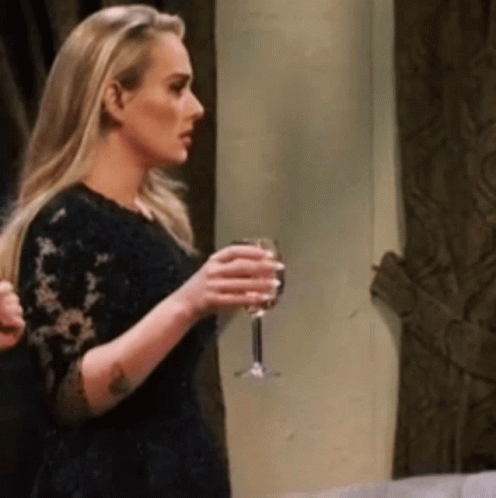 A Blonde Woman In A Black Dress Is Holding A Glass Of Wine . GIF