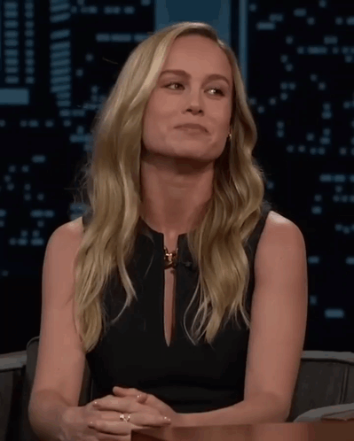 A Blonde Woman In A Black Dress Is Sitting At A Table GIF