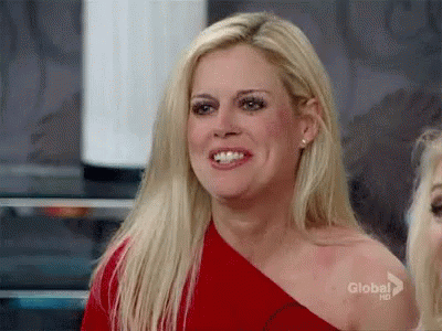 A Blonde Woman Is Wearing A Red One Shoulder Top And Smiling . GIF