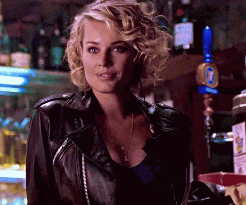 A Blonde Woman Wearing A Black Leather Jacket Stands In Front Of A Bar GIF