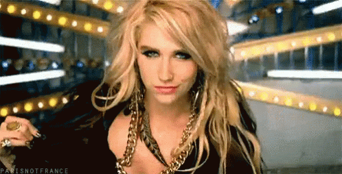 A Blonde Woman Wearing A Black Top And Gold Chains Looks At The Camera GIF