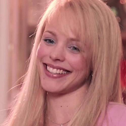 A Blonde Woman Wearing A Pink Shirt And A Necklace Is Smiling GIF