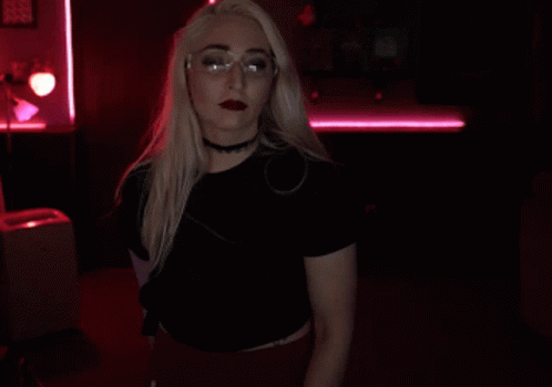 A Blonde Woman Wearing Glasses And A Choker Holds Her Finger To Her Lips GIF