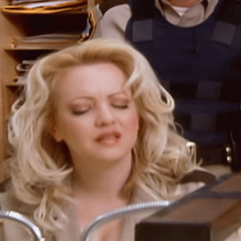 A Blonde Woman With Her Eyes Closed And A Man In A Blue Vest In The Background GIF