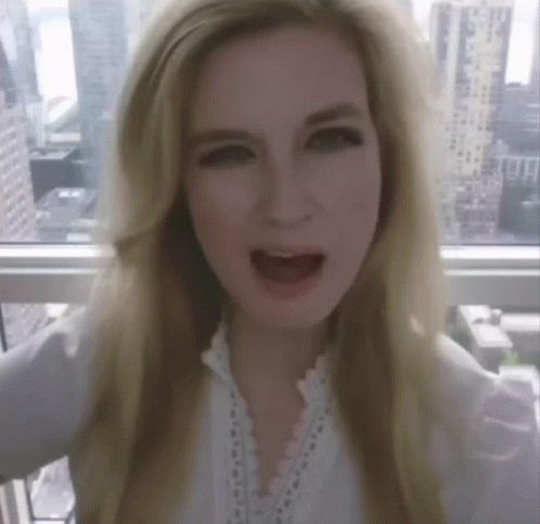 A Blonde Woman With Her Mouth Open Stands In Front Of A Window GIF