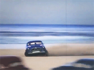 A Blue Car Is Driving Down A Sandy Beach Near The Ocean GIF