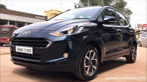 A Blue Hyundai Grand 110 Nios Is Parked In Front Of A Royal Hyundai Building GIF