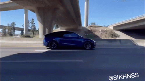 A Blue Tesla Model Y Is Driving Under A Highway Bridge GIF