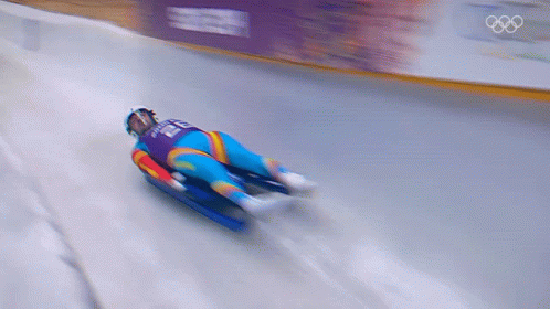 A Blurred Image Of A Person Sledding Down A Snowy Track With The Olympics Logo In The Background GIF