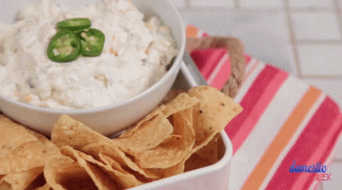 A Bowl Of Dip With Jalapenos On Top Next To A Bowl Of Chips GIF