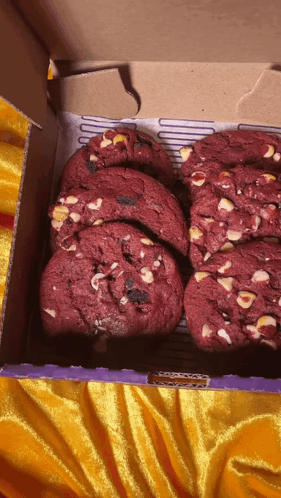 A Box Of Red Velvet Cookies With White Chocolate Chips Sits On A Yellow Cloth GIF