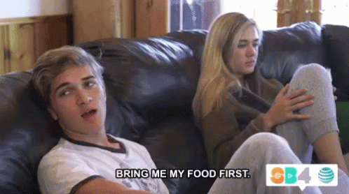 a boy and a girl are sitting on a couch and the girl is saying bring me my food first