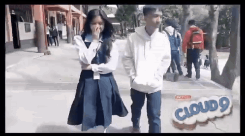 A Boy And A Girl Are Standing Next To Each Other On A Sidewalk . The Girl Is Covering Her Mouth . GIF