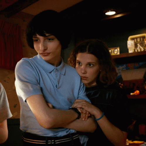 A Boy And A Girl Are Standing Next To Each Other With Their Arms Crossed In A Dark Room GIF
