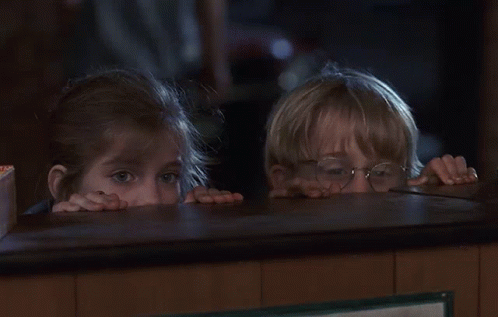 A Boy And A Girl Peeking Over A Counter In A Dark Room GIF