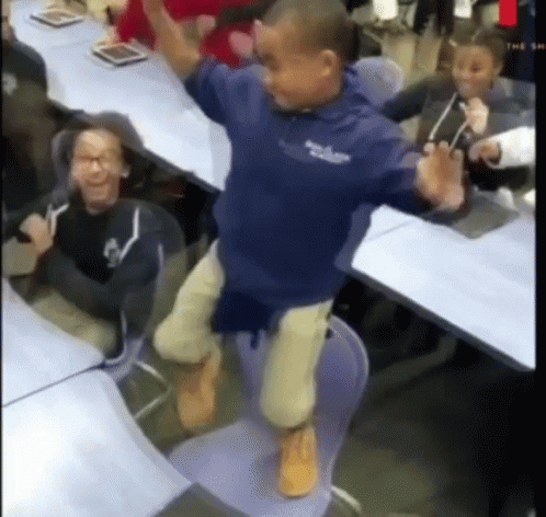 A Boy In A Blue Shirt Is Jumping In The Air While Sitting On A Chair In A Classroom . GIF