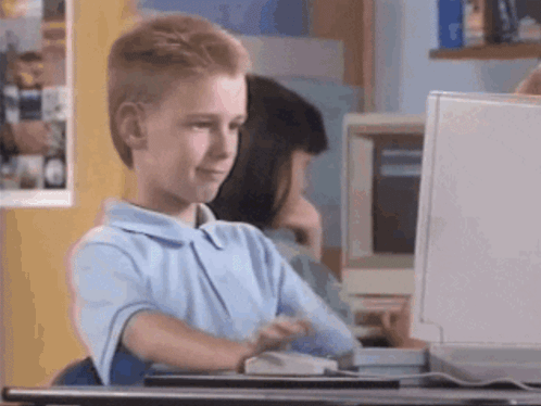 A Boy In A Blue Shirt Is Sitting At A Desk In Front Of A Computer Monitor . GIF