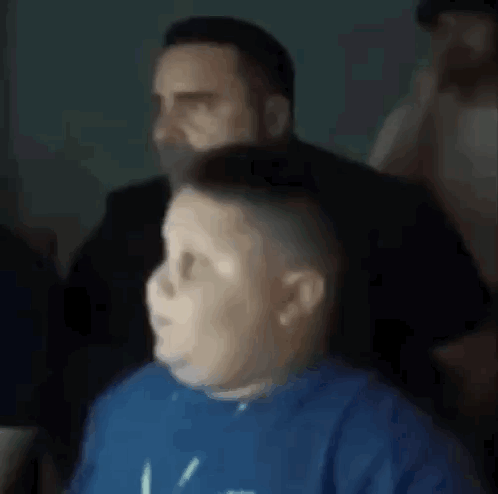 A Boy In A Blue Shirt Is Sitting In Front Of A Man In A Black Suit . GIF