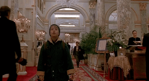 A Boy In A Green Coat Is Walking Down A Hallway In A Hotel GIF
