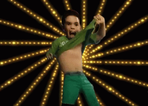 A Boy In A Green Shirt And Green Shorts Is Dancing In Front Of A Burst Of Lights GIF