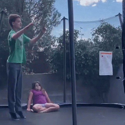 A Boy In A Green Shirt Is Jumping On A Trampoline While A Girl In A Pink Shirt Sits On The Ground . GIF