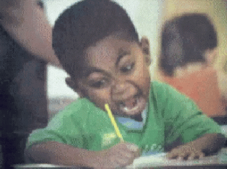 A Boy In A Green Shirt Is Writing With A Yellow Pencil In His Mouth . GIF