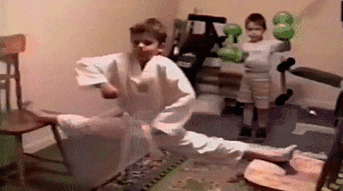 A Boy In A Karate Uniform Is Kicking A Chair While Another Boy Holds A Boxing Glove . GIF