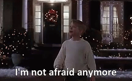 A Boy In A White Sweater Is Standing In Front Of A House With Christmas Lights On It . GIF