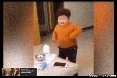 a boy in an orange sweater is standing in front of a table with a cell phone on it