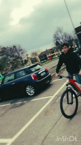 A Boy Is Riding A Bike In A Parking Lot With A Mini Cooper Behind Him GIF