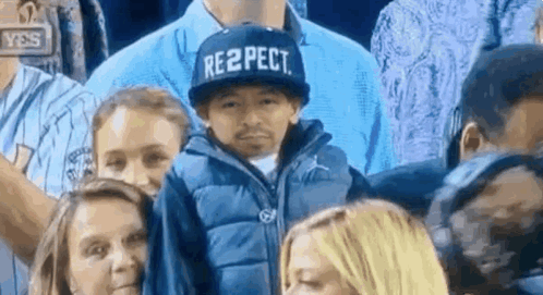 A Boy Wearing A Hat That Says Respect Is Sitting In A Crowd Of People . GIF