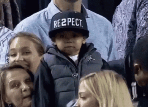 A Boy Wearing A Hat That Says Respect Is Sitting In A Crowd Of People . GIF
