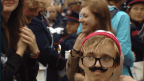 A Boy Wearing Glasses And A Fake Mustache Stands In A Crowd Of People With The Letters Nrk On The Bottom Right GIF