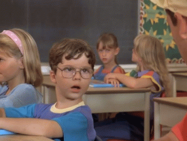 A Boy Wearing Glasses Sits At A Desk In A Classroom With Other Children GIF