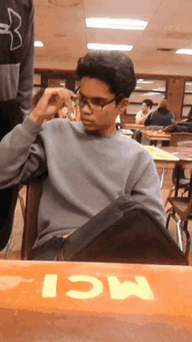 A Boy Wearing Glasses Sits At A Table With The Word Mci Written On It GIF