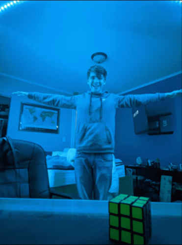 A Boy With His Arms Outstretched Is Standing Next To A Yellow Rubik 'S Cube GIF