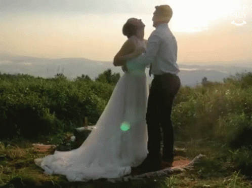 A Bride And Groom Are Dancing In A Field At Sunset . The Bride Is Wearing A Wedding Dress . GIF