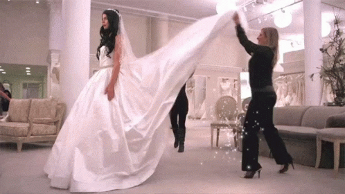 A Bride In A Wedding Dress Is Being Helped By A Woman GIF