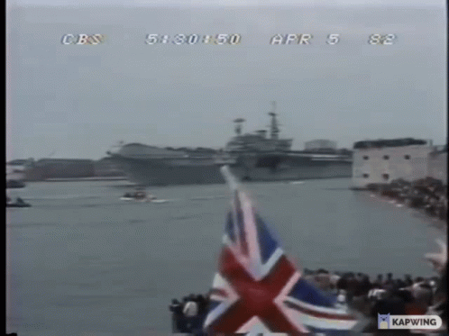A British Flag Is Flying In Front Of A Large Ship On The Water GIF