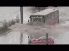 A Bus Is Driving Through A Flooded Street With A Car In The Water . GIF