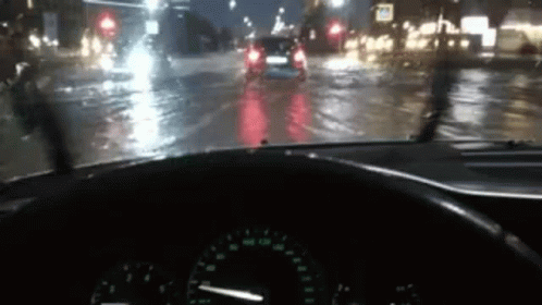 A Car Is Driving Down A Wet Street At Night With A Steering Wheel In The Foreground . GIF
