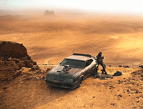 A Car Is Parked On A Dirt Hillside With A Man Standing Next To It And A Desert In The Background GIF