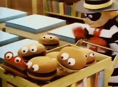 A Cartoon Character Is Standing Next To A Box Of Stuffed Hamburgers With Googly Eyes . GIF