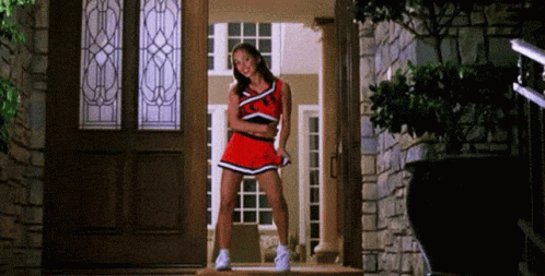 A Cheerleader Is Standing In Front Of A Door Wearing A Red Uniform . GIF