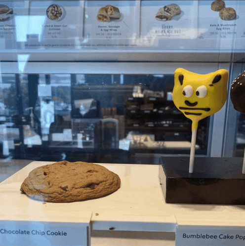 A Chocolate Chip Cookie And A Bumblebee Cake Pop Are On Display In A Bakery GIF
