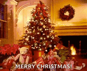A Christmas Tree Is Sitting In Front Of A Fireplace In A Living Room Surrounded By Presents And A Teddy Bear . GIF