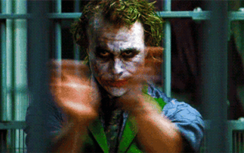 A Close Up Of A Joker Behind Bars With His Hands Outstretched GIF